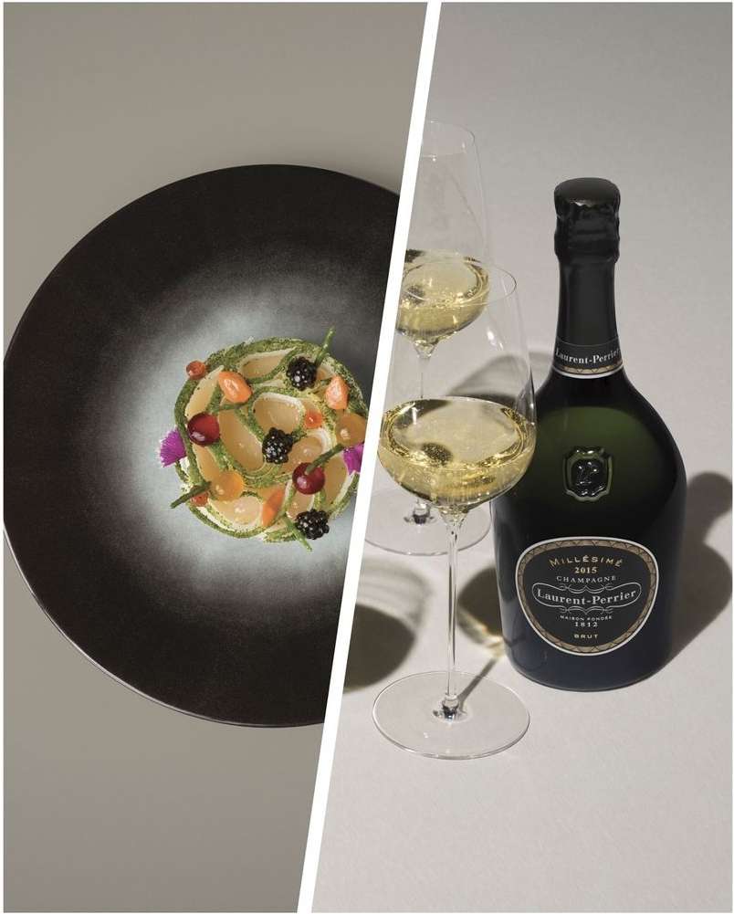 Still life featuring a plated salad with fruit and flowers next to champagne glasses and a bottle of Laurent-Perrier on a gray background.