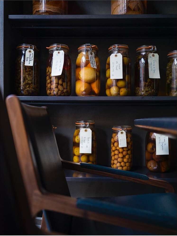 Display of preserved goods in glass jars on shelves, indoor setting with a foreground wood chair, dark and moody lighting.