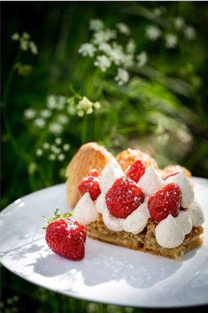 Close-up of a slice of mille-feuille with pastry cream and strawberries on a white plate, outdoors with a blurry green background.