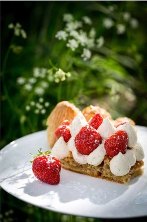 Close-up of a slice of mille-feuille with pastry cream and strawberries on a white plate, outdoors with a blurry green background.