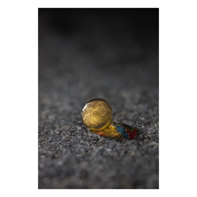 Close-up studio shot of a golden sphere with veined pattern, resting on a surface with paint streaks, against a blurred dark backdrop.