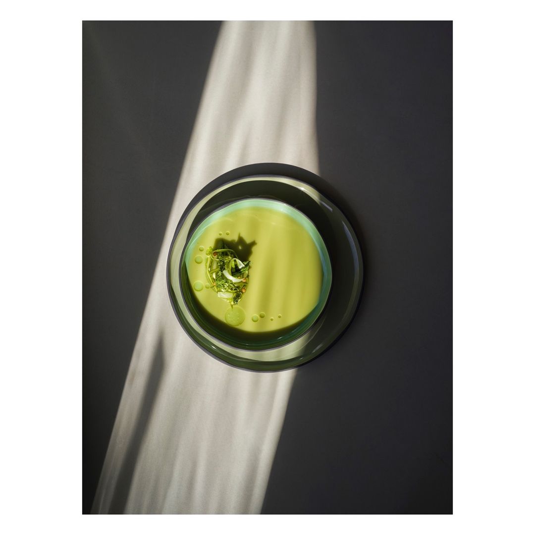 Overhead shot of light green soup with garnish in a glass bowl on a grey surface, partially illuminated by a beam of sunlight.