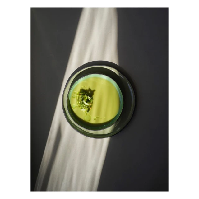 Overhead shot of light green soup with garnish in a glass bowl on a grey surface, partially illuminated by a beam of sunlight.