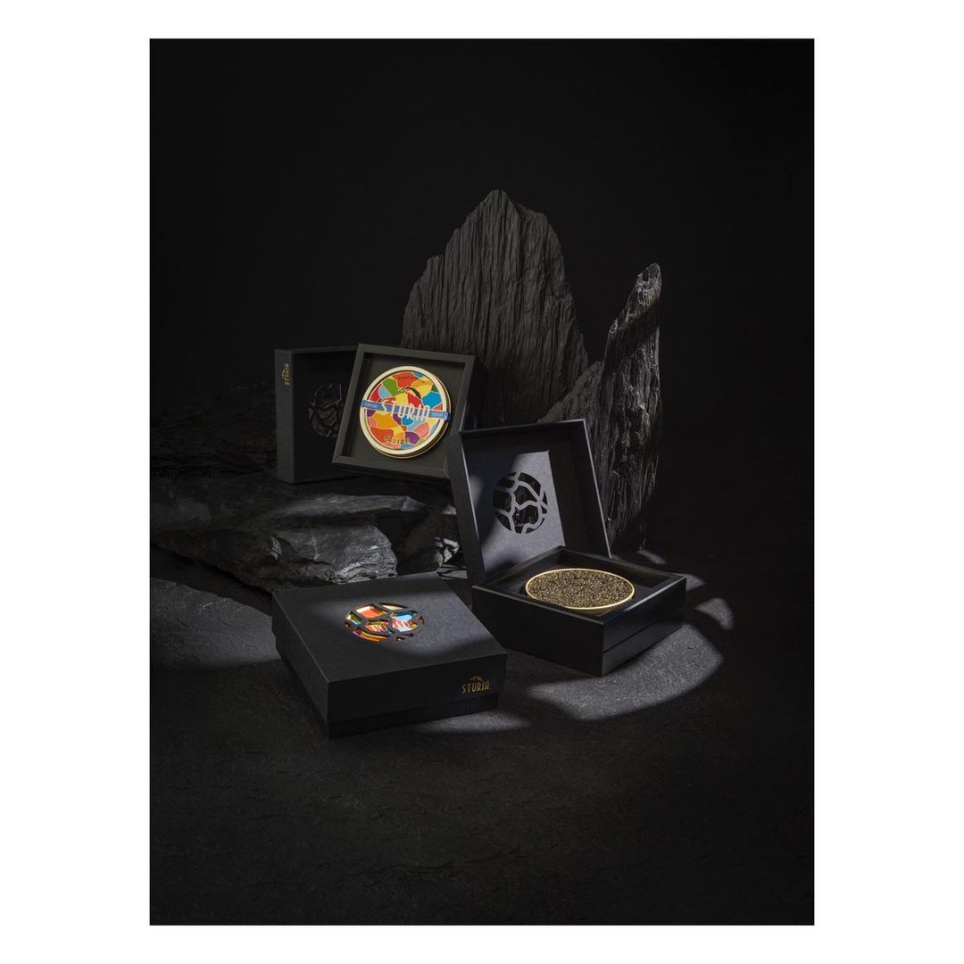 Four open black boxes display caviar, one featuring an abstract art lid, arranged against a dramatic black rock background.