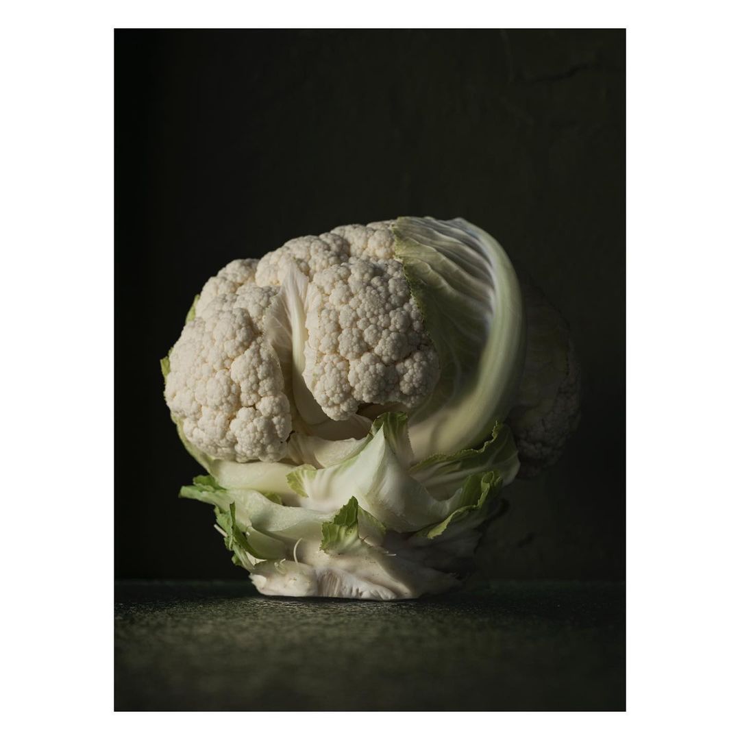 Close-up shot of a fresh cauliflower head with textured white florets and green leaves, set against a dark, moody background.
