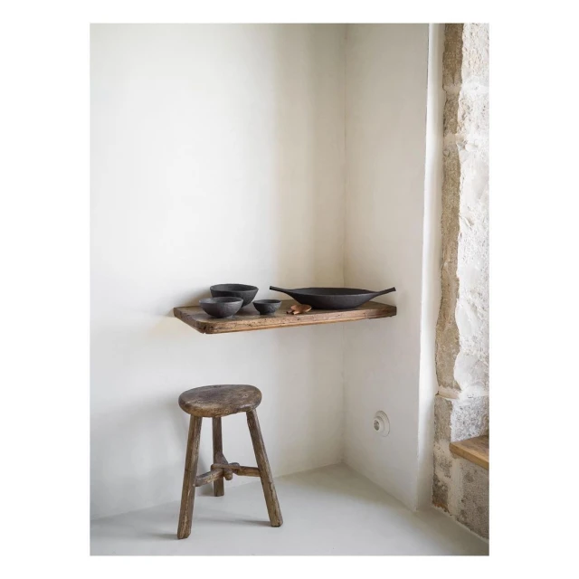 Rustic interior with wooden stool, shelf, and earthenware bowls against a white wall and stone column; minimalist design.