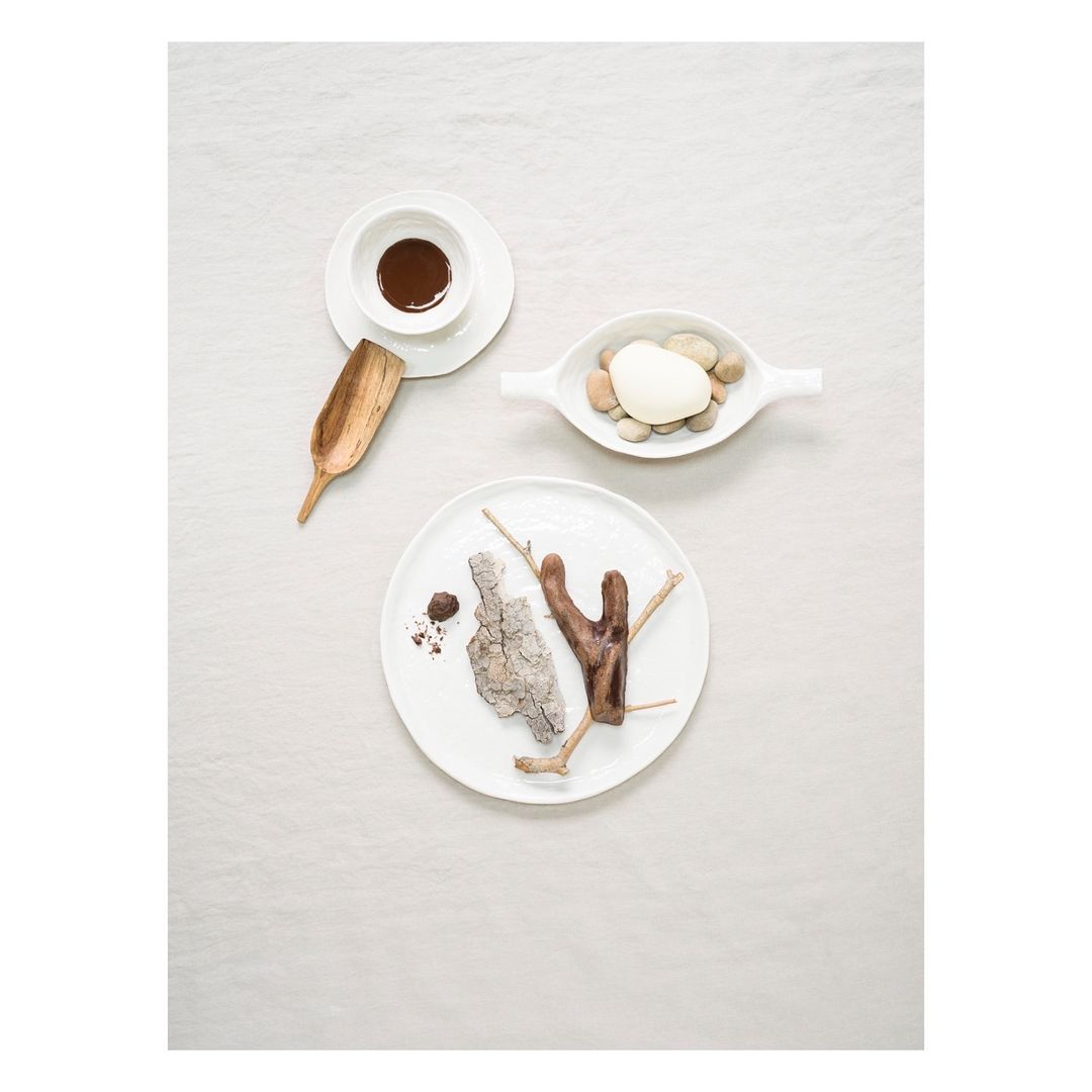 Overhead shot of tabletop arrangement with white plates, chocolate sauce, wooden scoop, pebbles, tree bark, and linen background.