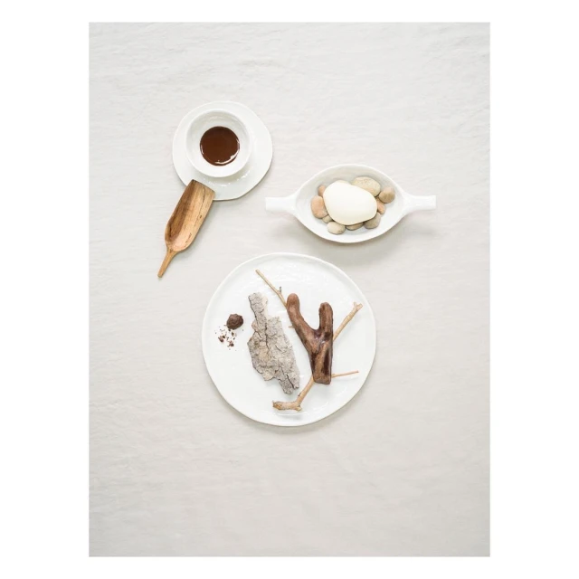 Overhead shot of tabletop arrangement with white plates, chocolate sauce, wooden scoop, pebbles, tree bark, and linen background.