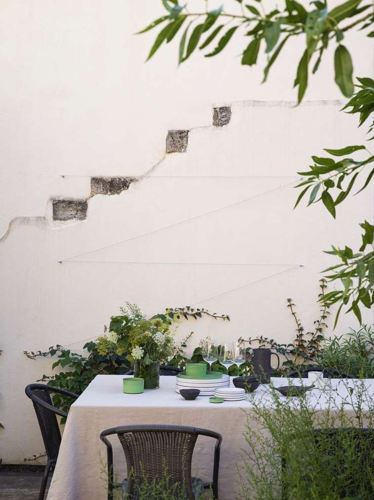 Outdoor dining scene features a table set for a meal, covered with a tablecloth, surrounded by chairs, with greenery in the background.