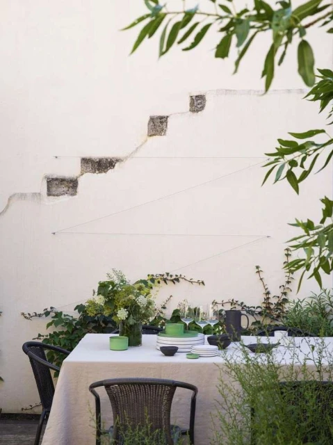 Outdoor dining scene features a table set for a meal, covered with a tablecloth, surrounded by chairs, with greenery in the background.