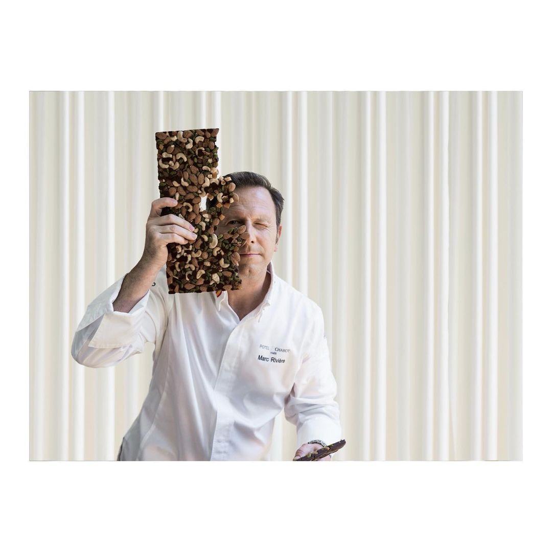 Chef in a white coat holds up a chocolate bar with nuts in front of a neutral background and looks at the camera with a slight smile.