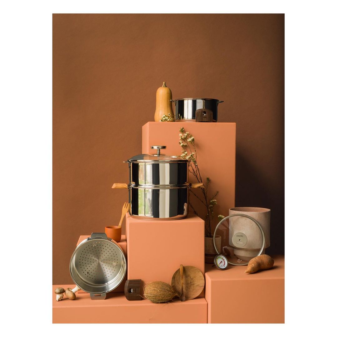 A variety of stainless steel cookware and produce arranged on peach-colored blocks against a tan background in a studio food shoot.
