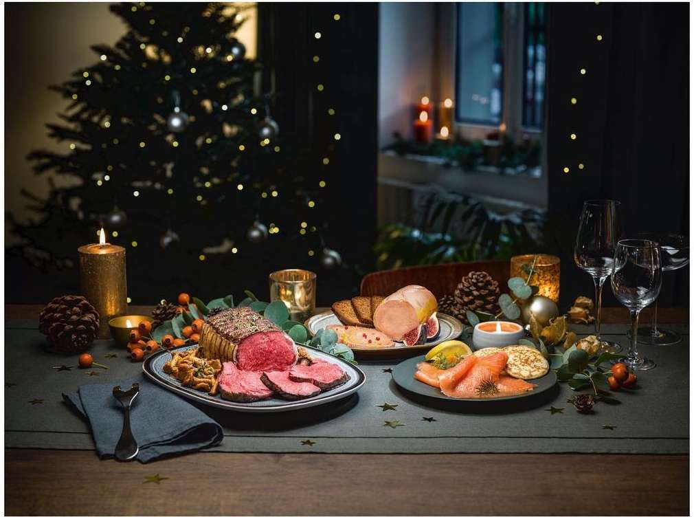 Festive holiday table setting indoors with roast beef, salmon, wine glasses, candles, and a Christmas tree in the background.