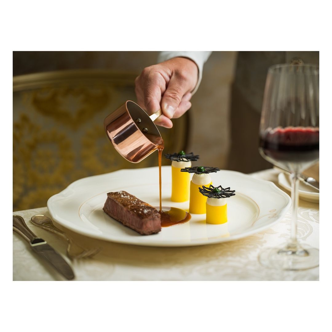 Chef pouring sauce over a steak dish with creative yellow cylinders, set on a fancy plate, accompanied by a glass of red wine.