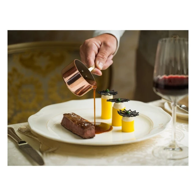 Chef pouring sauce over a steak dish with creative yellow cylinders, set on a fancy plate, accompanied by a glass of red wine.