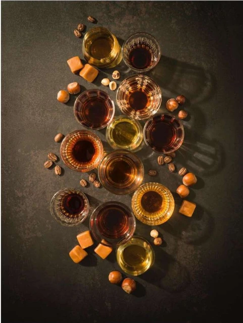 Overhead shot of arranged glassware with amber liquids, caramel candies, coffee beans, and hazelnuts on a dark, textured surface.