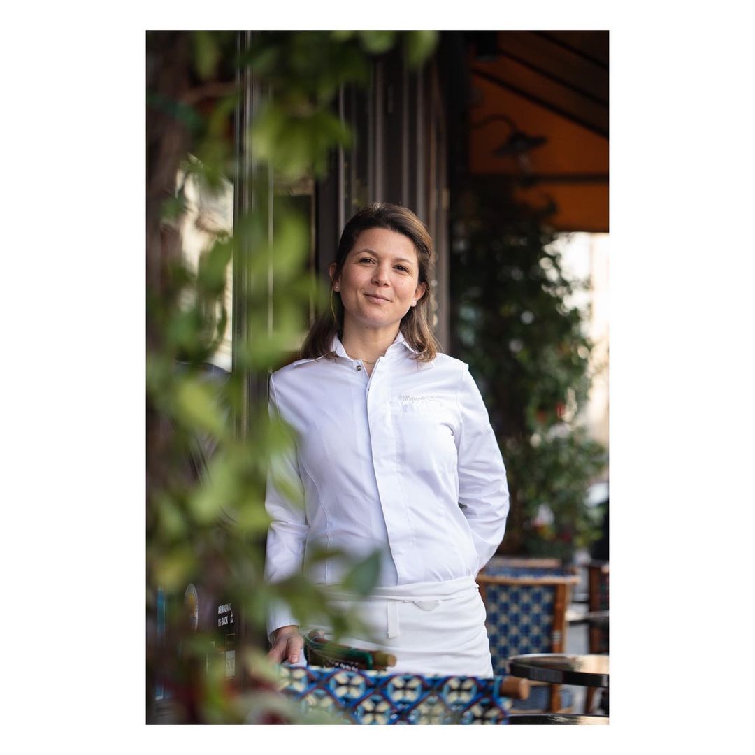 Portrait of an adult woman in a white chef's jacket outside a restaurant. She smiles with green plants visible in the frame.