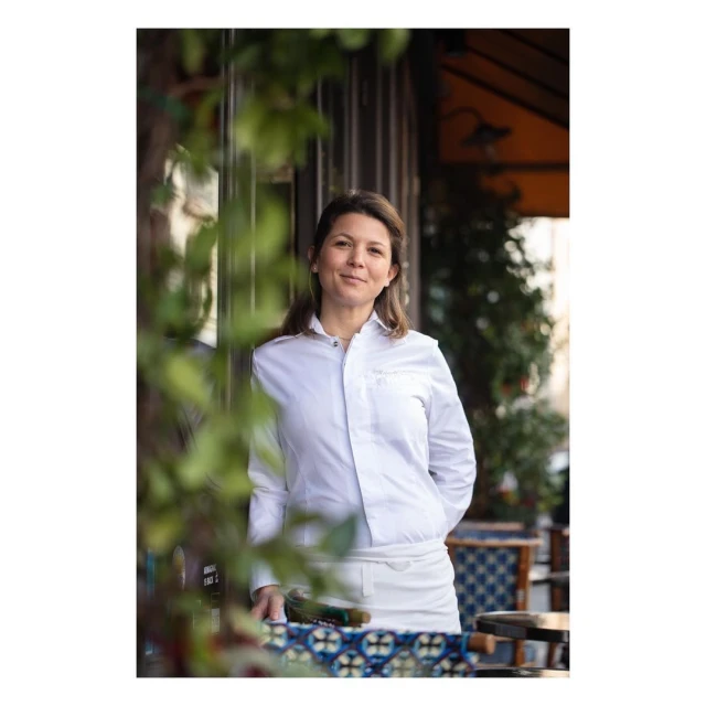Portrait of an adult woman in a white chef's jacket outside a restaurant. She smiles with green plants visible in the frame.