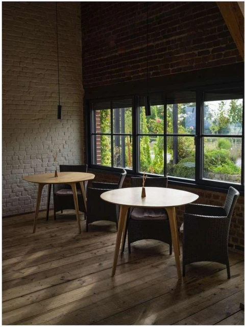 Interior cafe space featuring two round wooden tables with chairs, brick walls, a wood floor, and large windows overlooking greenery.