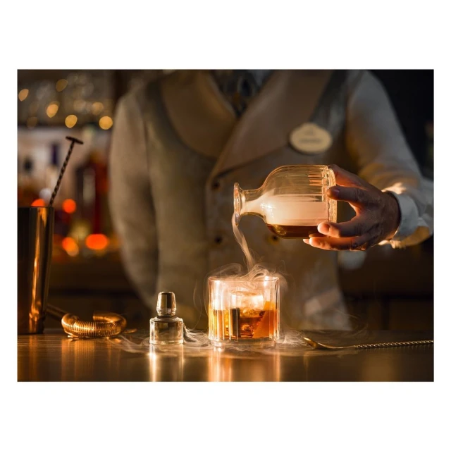 Bartender pours a cocktail from a glass decanter into a glass with ice, with a metal shaker and bar equipment on a wooden bar.