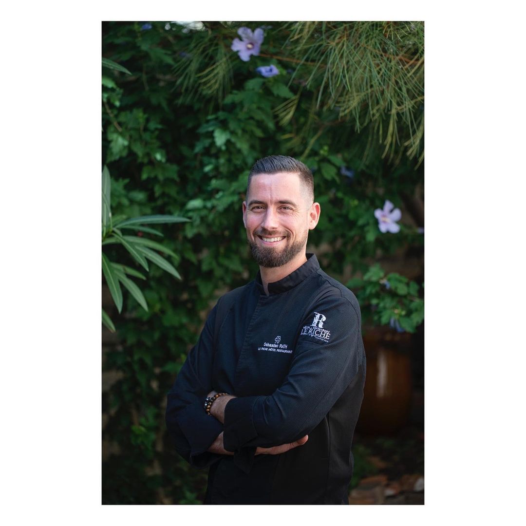 Close-up portrait of a smiling adult male chef in a black chef's coat, standing outdoors in front of green foliage and purple flowers.