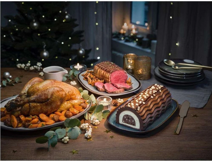 Holiday feast featuring roasted turkey, beef roast, and chocolate log cake arranged on a wooden table with festive holiday decorations.