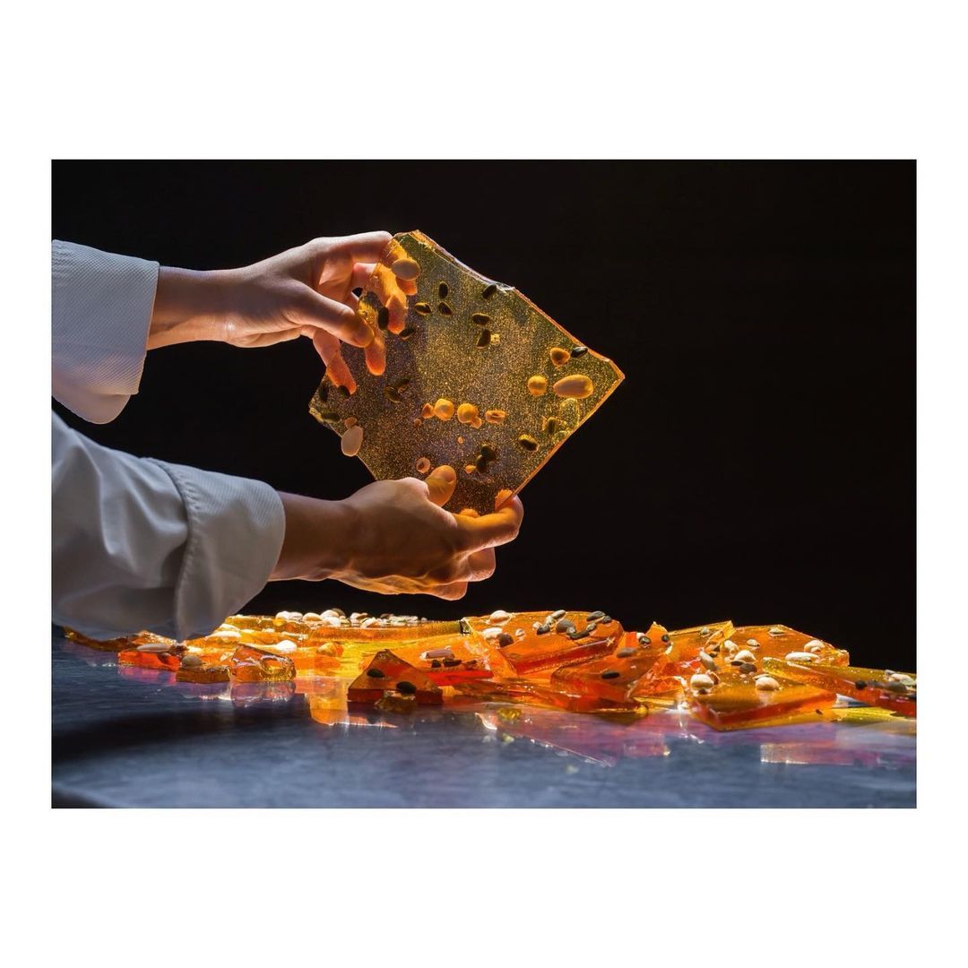 Close-up studio shot of chef's hands holding brittle candy with nuts, illuminated on a dark surface, showcasing culinary art.