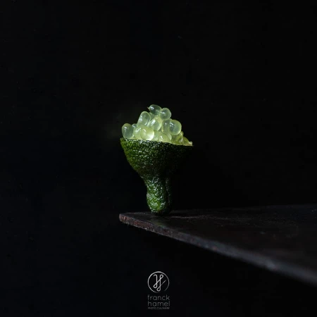 Close-up still life of translucent green finger lime vesicles mounded in a halved textured citrus peel on a dark surface