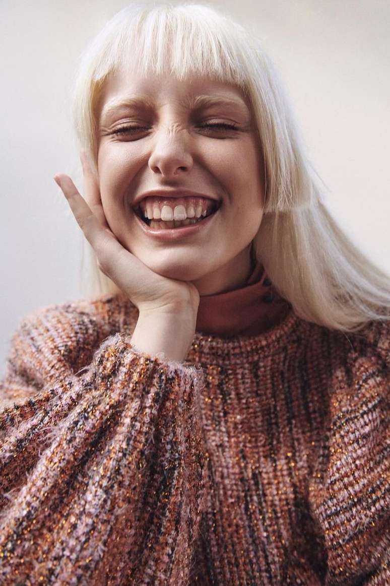 Close-up portrait of a smiling blonde woman with eyes closed, wearing a patterned sweater, set against a plain background in soft light.