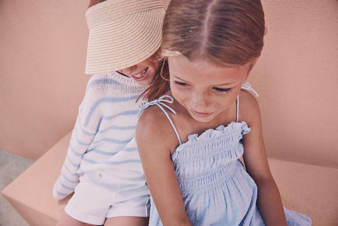 Two young girls in light-colored summer clothing, one wearing a sun visor, sitting in an indoor studio setting with soft, diffused lighting.