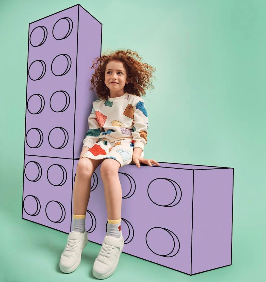 A young girl with curly red hair is sitting on purple toy blocks in a kids' fashion studio with a mint green background.