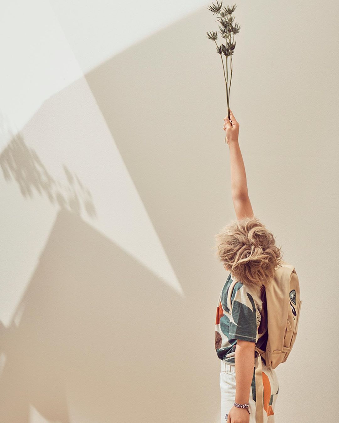 Child stands with back to camera holding flowers, wearing patterned shirt and backpack against white wall with light and shadow.