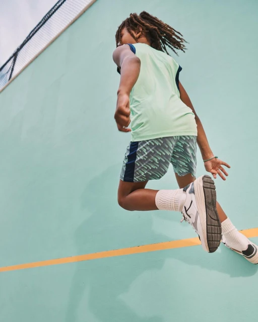 Action shot of a child with dreadlocks jumping against a teal wall, wearing a light green athletic shirt, patterned shorts, white socks, and sneakers.