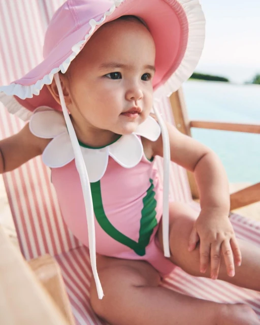 A baby in a pink swimsuit and sun hat sits on a striped beach chair with a serene expression, under bright natural light outdoors.