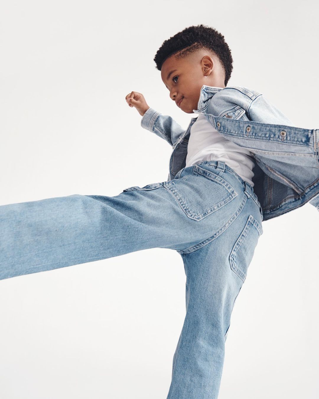 A boy in light blue denim jacket and jeans strikes a playful pose against a white backdrop, showcasing youthful energy and style.