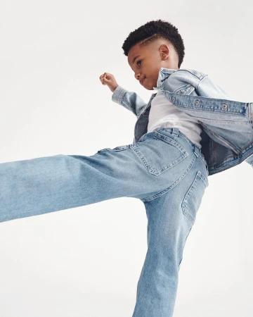 A boy in light blue denim jacket and jeans strikes a playful pose against a white backdrop, showcasing youthful energy and style.