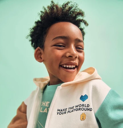 A smiling young boy with curly hair wears a light-colored zip-up hoodie with Lego-themed designs against a light green backdrop.