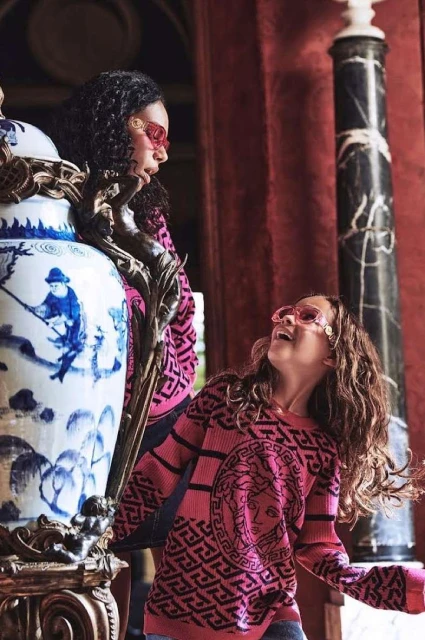 Two young female fashion models pose indoors in pink Versace sweaters with sunglasses, next to a large ornate vase and marble columns.