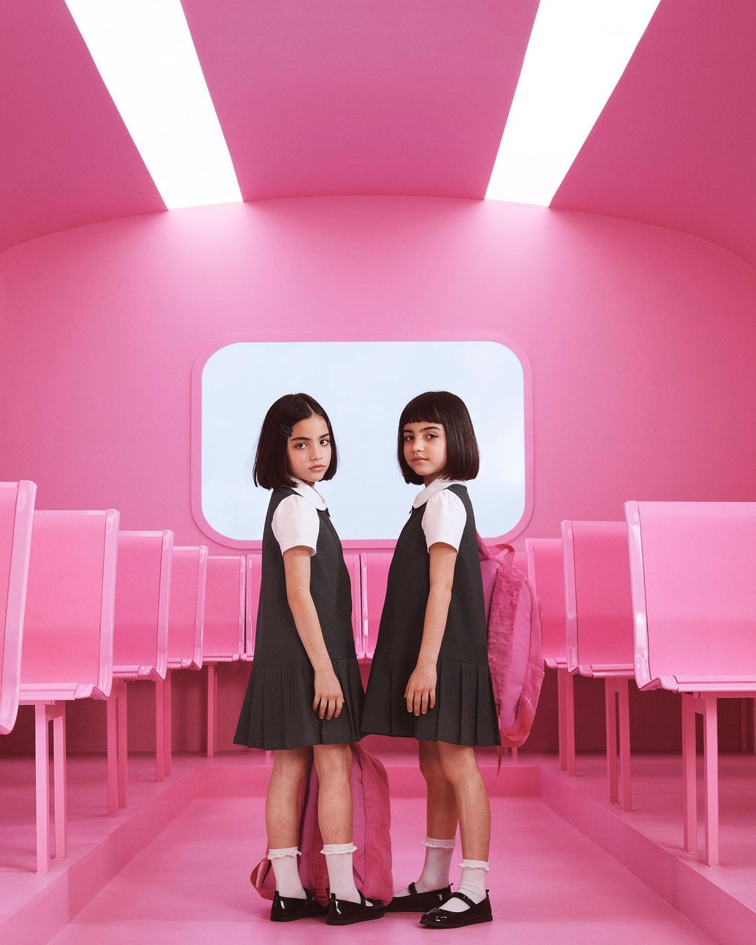 Two young girls in school uniforms stand in a pink hallway, each with a pink backpack. Lit by fluorescent lights in an indoor environment.
