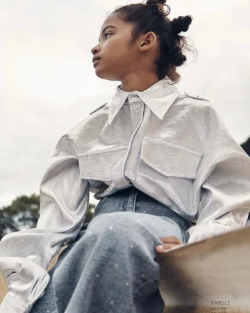 Young girl poses in a silver, collared shirt and blue skirt, looking up and away from the camera in an outdoor setting.