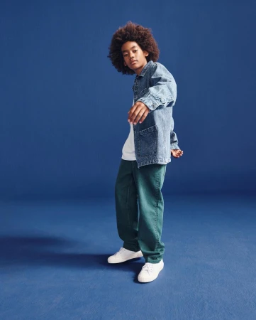 Young boy with curly hair wearing a denim jacket and green pants against a blue background