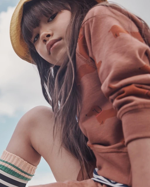 Close-up of a child model wearing a brown patterned sweatshirt and a yellow straw hat against a light blue sky.