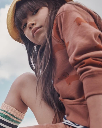 Close-up of a child model wearing a brown patterned sweatshirt and a yellow straw hat against a light blue sky.