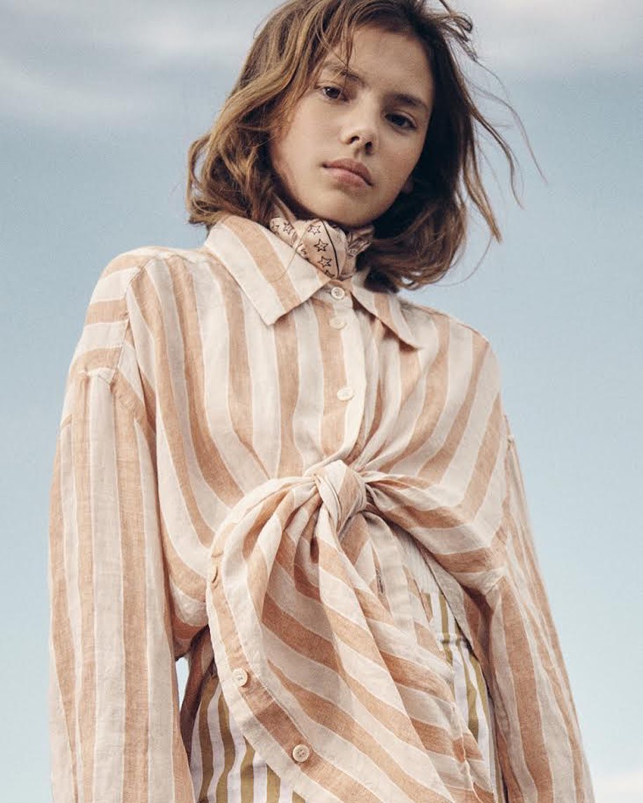 Young woman in a striped button-down shirt and neck scarf photographed from a low angle against a clear sky.
