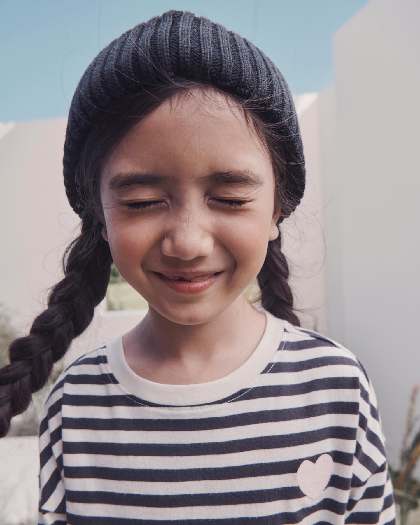 Portrait of a young girl with a beanie and braids, smiling with her eyes closed, wearing a striped shirt with a heart detail, outdoors.