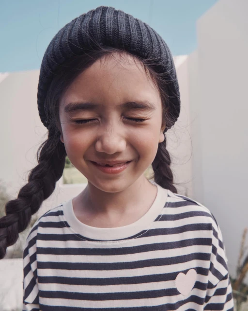 Portrait of a young girl with a beanie and braids, smiling with her eyes closed, wearing a striped shirt with a heart detail, outdoors.