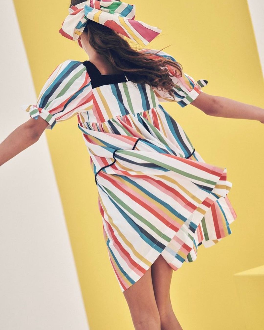 Girl wearing a colorful striped dress and matching headscarf twirls in front of a yellow and white backdrop in a studio setting.