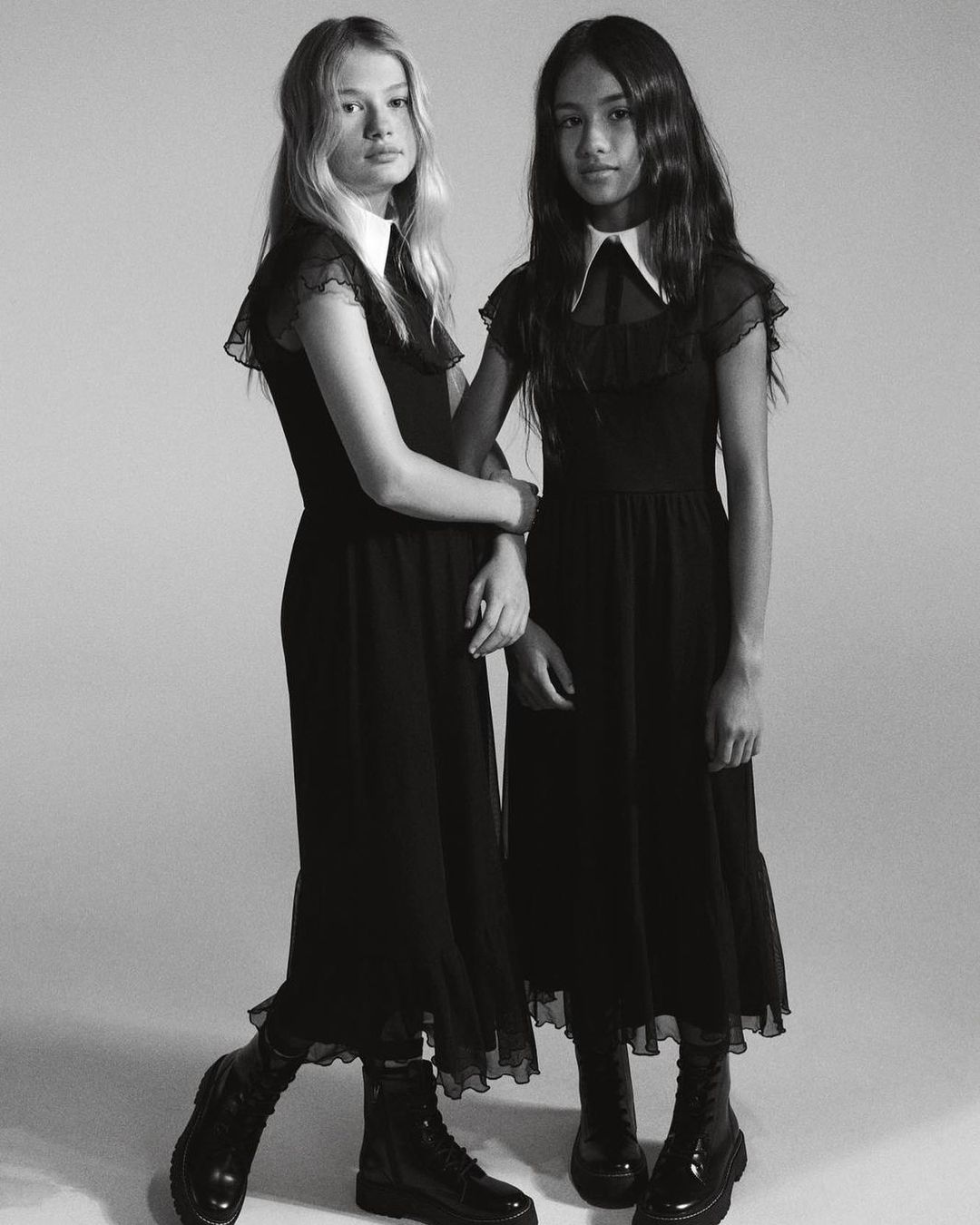 Two young women stand arm-in-arm in a black and white portrait, wearing matching black dresses with white collars and black boots.