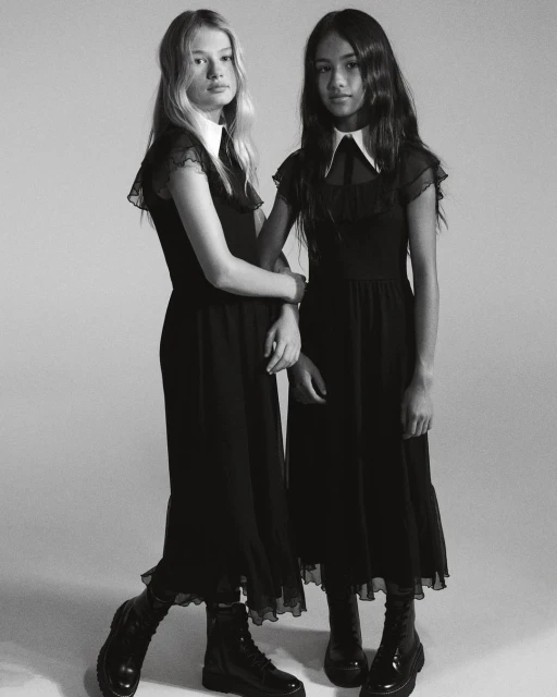 Two young women stand arm-in-arm in a black and white portrait, wearing matching black dresses with white collars and black boots.