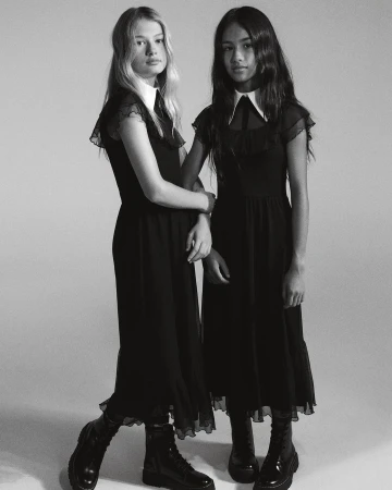 Two young women stand arm-in-arm in a black and white portrait, wearing matching black dresses with white collars and black boots.